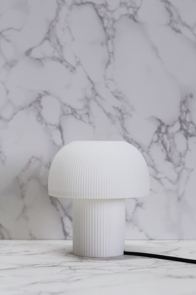 A table lamp with a white, rounded silhouette and a gently domed top, resembling a mushroom, with a base available in cream white and charcoal black, placed on a marble surface against a marble background.